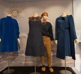 Conservator stands between three mannequins displaying blue garments from Princess Grace Kelly’s wardrobe, including a beaded-neckline dress and tailored coats, during gallery installation.