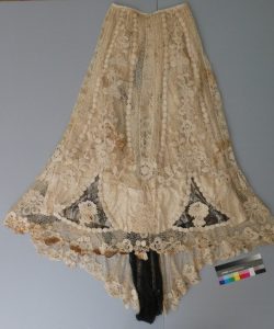 Full view of a historic cream-colored lace skirt with floral motifs, showing areas of discoloration, staining, and structural loss along the hem and panels.